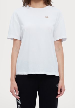 Žena oblečená v jednoduchém bílém tričku s krátkým rukávem a malým zlatým logem EA7 na hrudi a v černých kalhotách s bílým nápisem EA7.