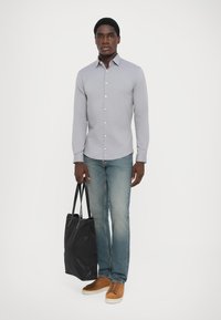 Lys grå skjorte med knapp, denim jeans, brune sneakers og en svart tote-bag. Skjorten har en skreddersydd passform og krage.