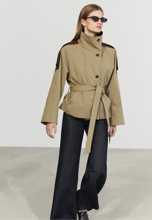 Donna che indossa una giacca beige con cintura e bottoni neri, pantaloni scuri a gamba larga, scarpe nere e piccoli occhiali da sole neri, in piedi su un pavimento semplice.