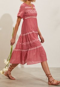 Vaaleanpunainen kevyt mekko, jossa on lyhyet hihat, kerroksellinen design, brodeeratut yksityiskohdat ja pyöreä kaula-aukko. Malli kantaa vaaleanpunaisia tulppaaneja.