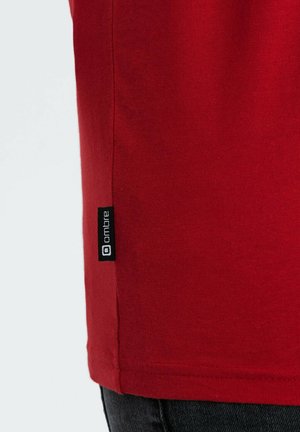 T-shirt en coton rouge avec une texture lisse, présentant une étiquette noire sur le côté avec "ombre" imprimé. Design à manches courtes.