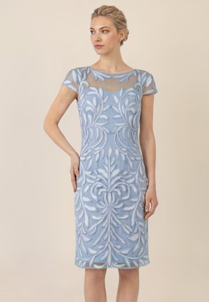 Frau trägt ein knieumspielendes hellblaues Kleid mit weißer Blumenstickerei und transparenten kurzen Ärmeln, steht mit einer Hand in der Hüfte.