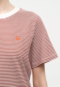 Κοντινό πλάνο ενός ατόμου που φοράει μια κόκκινη και λευκή ριγέ μπλούζα με μια μικρή πορτοκαλί καρδιά κεντημένη στο στήθος.