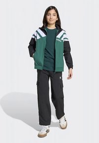 Πράσινο και μαύρο μπουφάν με λευκές ρίγες, φορεμένο πάνω από πράσινο T-shirt, σε συνδυασμό με μαύρο παντελόνι cargo και λευκά αθλητικά παπούτσια.