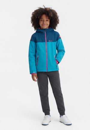 Lokkis juustega laps, kes kannab sinist ja türkiissinist kapuutsiga jopet, tumedat halli pükse ja valgeid tenniseid, seistes ühe käe taskus.