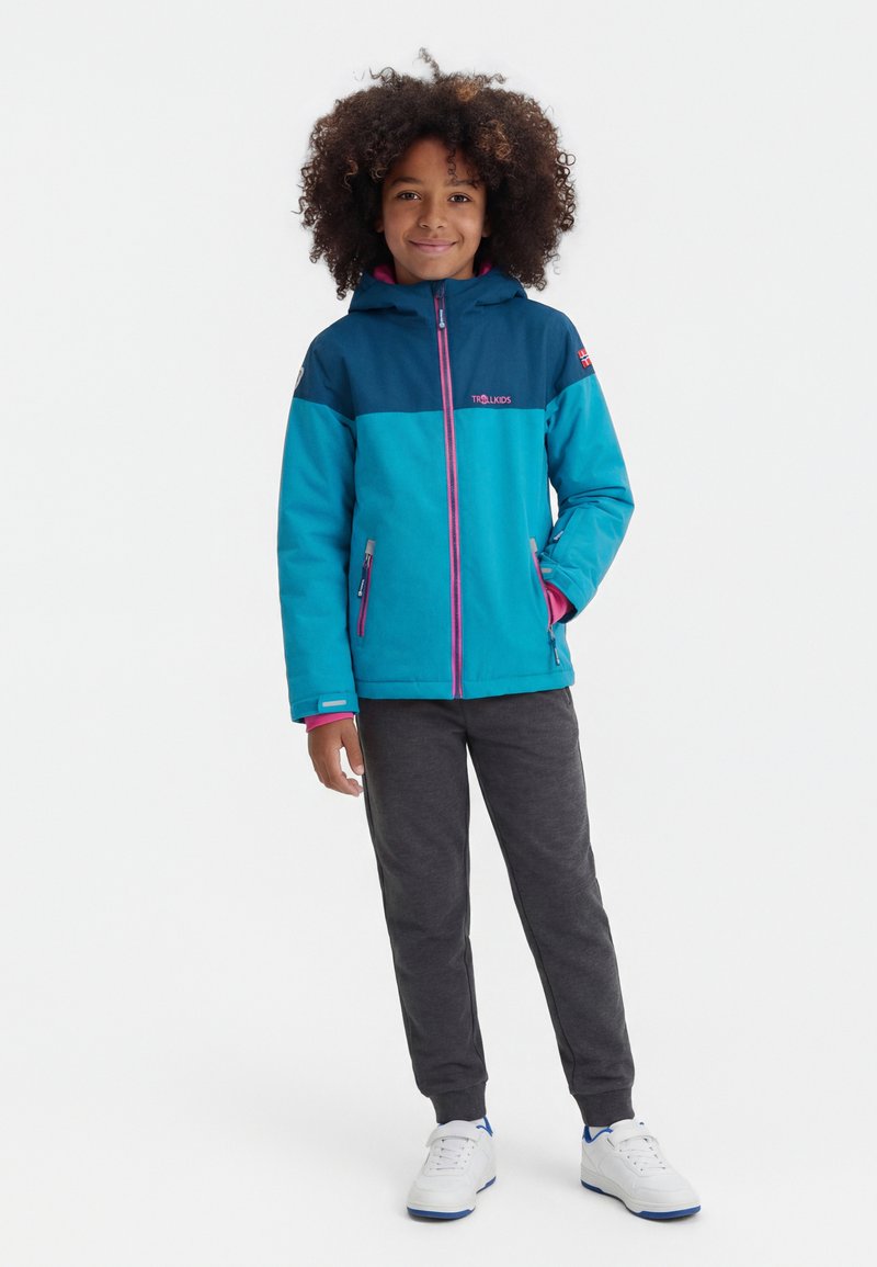 Kind met krullend haar, gekleed in een blauwe en turquoise capuchonjas, donkergrijze broek en witte sneakers, staand met één hand in de zak.