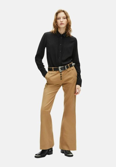 Zwarte button-up shirt met een getextureerde voorkant; tan wijde broek met voorplooien en vijf knopen; gedragen met een zwarte riem en laarzen.