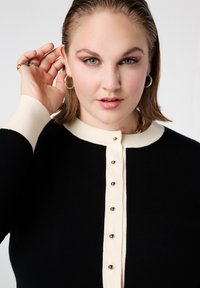 Zwart-witte cardigan met een knoopsluiting aan de voorkant, voorzien van goudkleurige knopen en contrasterende geribbelde manchetten. Zacht gebreid materiaal en een aangesloten pasvorm.