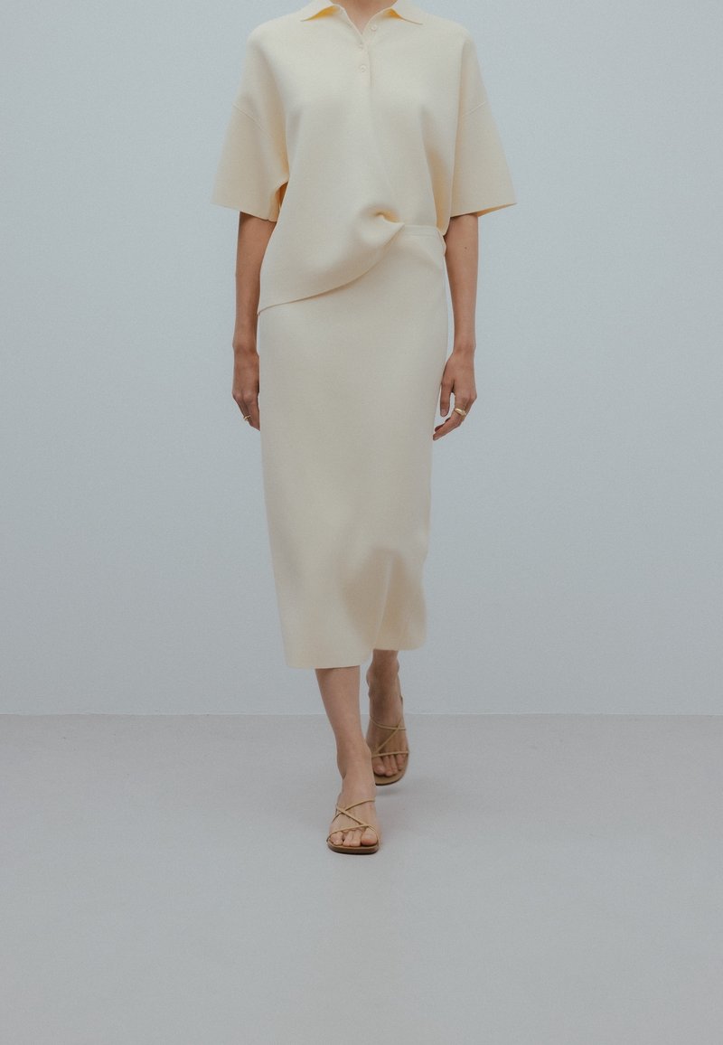 Personne marchant avec un haut ample beige et une jupe midi assortie, portant des sandales beiges à lanières sur un sol et un mur gris unis.
