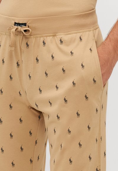 Beige színű melegítő nadrág, zsinóros derékkal, ismétlődő fekete póló játékos logó mintával, puha anyagból készült.