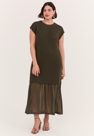 Vrouw draagt een mouwloze donkergroene olijfkleurige gebreide jurk met een doorschijnende olijfgroene rokkenlaag en hoge hakken met slangprint, staand tegen een effen achtergrond.