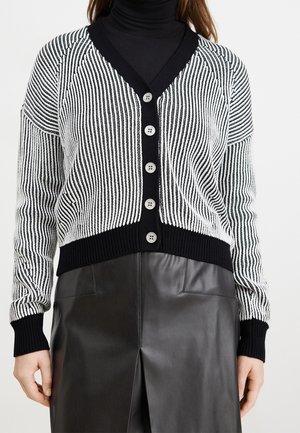 Femme portant un cardigan rayé noir et blanc boutonné sur un col roulé noir, associé à une jupe en cuir noire avec une fente devant.