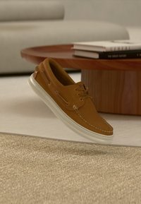 Καφέ δερμάτινο παπούτσι τύπου boat με λευκή σόλα από καουτσούκ, κορδόνια και λεπτομέρειες ραφής. Κρεμασμένο πάνω από μια υφασμάτινη μπεζ επιφάνεια.