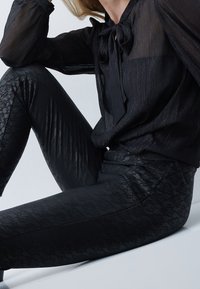 Zwarte leggings met kantpatroon, gecombineerd met een doorschijnend zwart blouse met een striknek. De outfit heeft een combinatie van soepele texturen en een aansluitend ontwerp.
