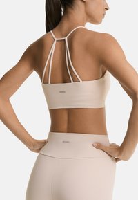 Beige sports bra with thin, crisscross straps and logo at the back; paired with matching high-waisted leggings in a smooth, stretchy fabric.