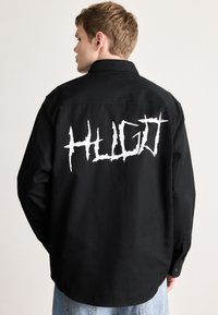 Μαύρο πουκάμισο με μυτερό γιακά, μακριά μανίκια και μεγάλο λευκό γραφικό λογότυπο "HUGO" στην πλάτη. Ομαλό ύφασμα, άνετη εφαρμογή.