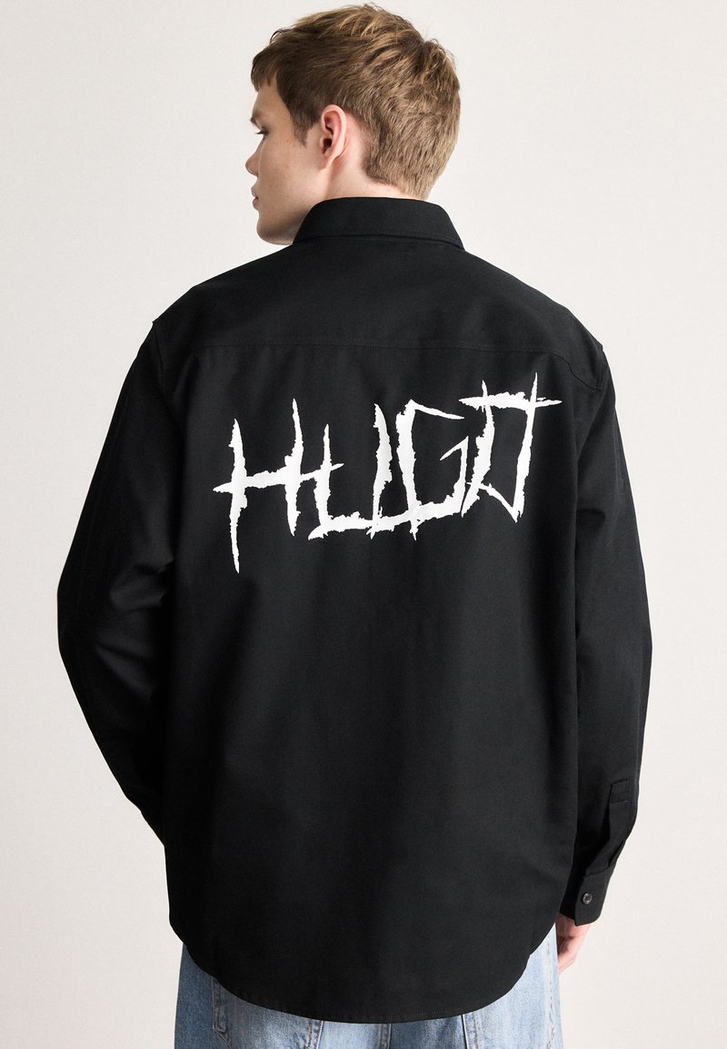 Μαύρο πουκάμισο με μυτερό γιακά, μακριά μανίκια και μεγάλο λευκό γραφικό λογότυπο "HUGO" στην πλάτη. Ομαλό ύφασμα, άνετη εφαρμογή.