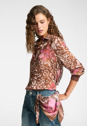 Mujer con cabello rizado que lleva una blusa estampada de flores en tonos marrón y rosa, jeans azules y una bufanda a juego atada a la cintura.