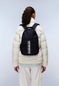 Marinblå ryggsäck med en slät yta, utrustad med en logotyp och sidofickor i nät, bärs över en vit puffjakke och en ljusrandig skjorta.