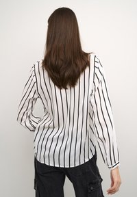 Valge pika varrukaga blouse, millel on vertikaalsed mustad triibud, ümmargune äär ja nööbiga mansetid, koos tumehallide cargo pükstega.