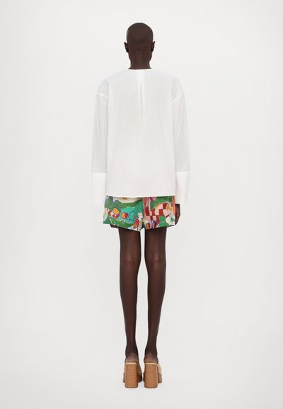 Witte lange mouwen shirt met verticale strepen en brede manchetten, gecombineerd met kleurrijke shorts met patronen in groen, rood en blauw.