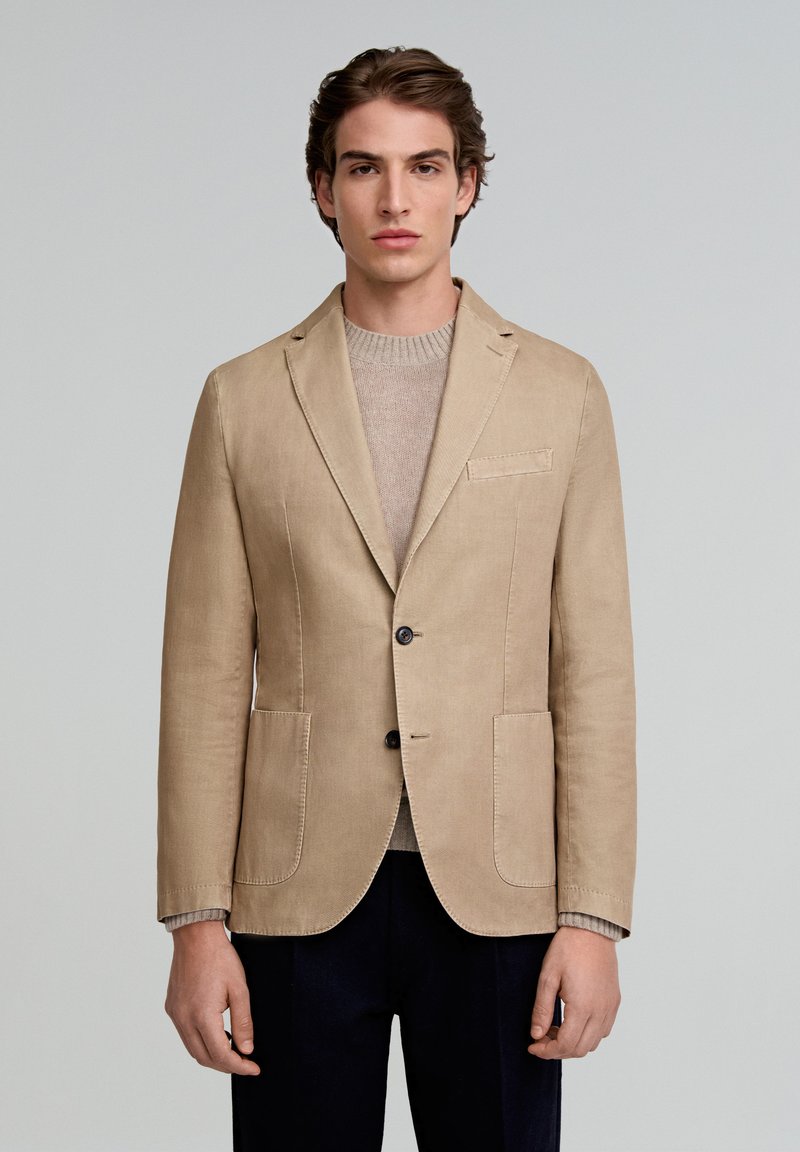 Blazer de lino beige con dos bolsillos frontales, dos botones y solapas con muesca, llevado sobre un suéter gris claro, combinado con pantalones oscuros.