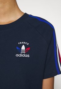 Camiseta color navy con un logo "adidas" bordado en blanco y colores de la bandera francesa. Detalles a rayas en rojo, blanco y azul a lo largo de los hombros.