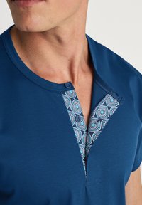 Blauwe shirt met korte mouwen, voorzien van een patroon met geometrische vormen op de knoopsluiting, twee blauwe knopen en een gladde, zachte textuur.