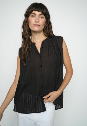 Pikkade laineliste juustega naine, kes kannab musta varrukateta nööbitavat pluusi ja valgeid pükse, seisab ühevärvilise heleda tausta ees.