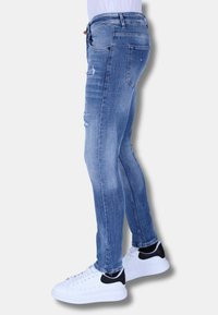 Slim-fit blauwe denim jeans met versleten details, lichte wassing en vijf-pocketontwerp, gecombineerd met witte sneakers met zwarte accenten.