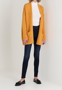Jersey de color mostaza con puños largos y bolsillos frontales, llevado sobre un suéter de cuello alto blanco y combinado con jeans ajustados oscuros y zapatos negros.
