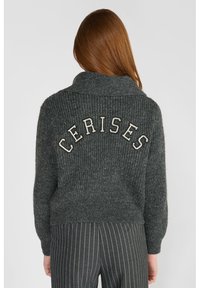 Pull en-maille gris avec un col châle, présentant "CERISES" brodé en blanc au dos. Porté avec un pantalon noir à rayures.