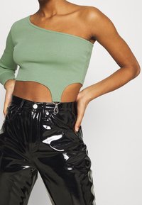 Frau in einem grünen Ein-Schulter-Crop-Top mit Frontausschnitt und schwarzen, glänzenden, hochgeschnittenen Hosen, die Hände auf den Hüften.