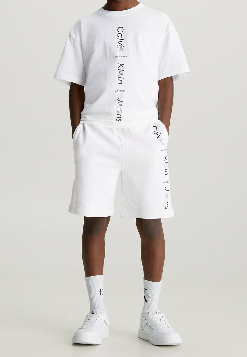 Witte katoenen t-shirt en korte broek set met verticale zwarte tekstbranding. Inclusief zijzakken en een ronde halslijn, gecombineerd met witte sneakers.