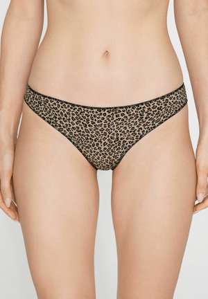 Femme portant un sous-vêtement style bikini imprimé léopard, montrant le ventre et le haut des cuisses sur un fond uni.