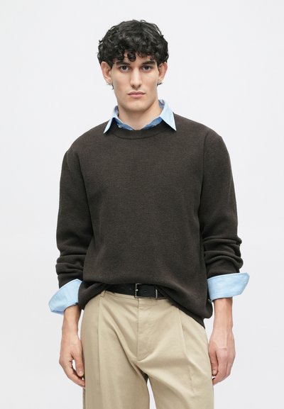 Καφέ πλεχτό πουλόβερ με ραβδωτή υφή, φορεμένο πάνω από ένα ανοιχτό μπλε πουκαμισάκι με γιακά. Το παντελόνι είναι μπεζ, με μαύρη ζώνη και χαλαρή εφαρμογή.