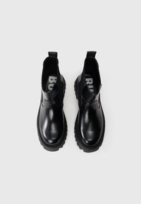 Botas de tornozelo pretas com acabamento brilhante, painéis laterais elásticos, alça de puxar e sola de borracha grossa. Apresentam bico redondo e detalhamentos de costura mínimos.