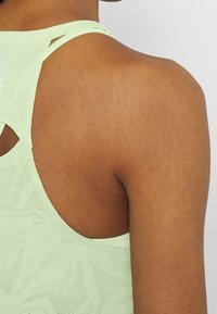 Detailní záběr na horní část zad a rameno osoby oblečené v lehkém zeleném sportovním topu s vzorovaným, ventilačním designem tkaniny.