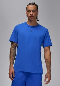 T-shirt blu a girocollo in cotone con maniche corte, caratterizzata da un logo Jordan nero sul petto. Il modello ha capelli intrecciati e tatuaggi visibili.