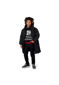 Svart dunjakke, grå genser med striper, røde detaljer, svarte jeans og svarte hodetelefoner. Joggesko med hvite såler. Avslappet antrekk.