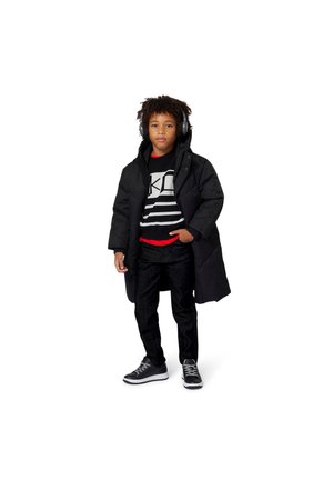 Juodas pūkinis paltas, pilkas džemperis su juostelėmis, raudonas akcentas, juodos džinsai ir juodos ausinės. Sportbačiai su baltais padais. Kasdienis aprangos stilius.