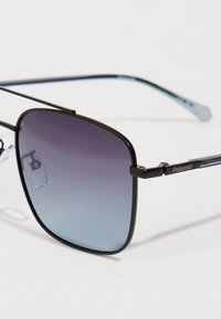 Mustat metallikehyksiset Polaroid aurinkolasit, joissa on suuret neliömäiset sinivioletit liukuvärioptiikat ja säädettävät nenätyynyt valkoista taustaa vasten.