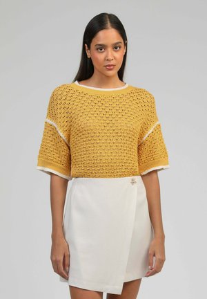 Femme portant un pull en maille jaune moutarde à manches courtes et une jupe portefeuille blanche avec une petite broche décorative.
