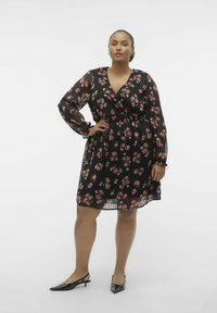 Robe portefeuille noire à motifs floraux avec de longues manches transparentes, dotée d'une taille ceinturée et de motifs de roses roses. Portée avec des escarpins noirs à bout pointu.