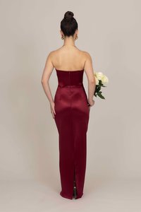 Frau mit einem Haarknoten, die ein trägerloses rotes Satinkleid trägt, während sie weiße Rosen hinter ihrem Rücken hält und vor einem schlichten Hintergrund steht.