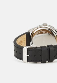 Montre ronde en argent avec un bracelet en cuir noir, présentant une texture lisse, une boucle en métal et un logo gravé sur le bracelet.