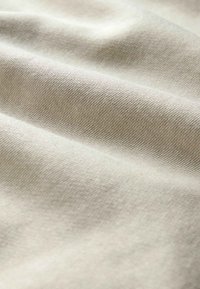 Světlé béžové plátno s jemnou texturou a nenápadným vzorem tkaní, se zlehka složenými vrstvami vytvářejícími hloubku a rozměr.