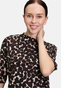 Kobieta o jasnej karnacji i niebieskich oczach, uśmiechająca się i dotykająca szyi, ubrana w czarną i brązową bluzkę w panterkę z rękawami do łokcia.