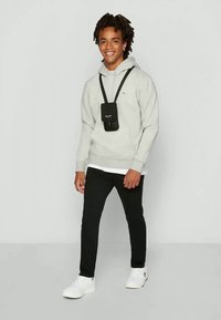 Jovem sorridente, vestindo um hoodie cinza claro, bolsa transversal preta, calças pretas e sapatilhas brancas, posando contra um fundo claro e liso.