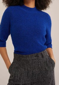 Maglione blu lavorato a maglia con collo rotondo e maniche corte, abbinato a pantaloni grigi con texture caratterizzati da un dettaglio con bottone e tasche laterali.
